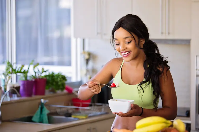 A happy, fit young woman in her kitchen, eating fruit and cereal.