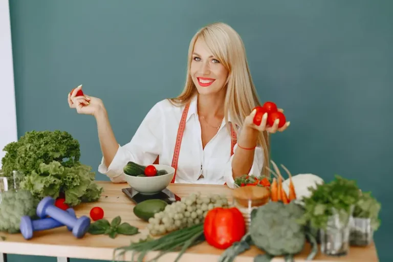 A smiling, slim, blonde, healthy looking woman sitting on a chair behind a table with various vegetables and greens on it.